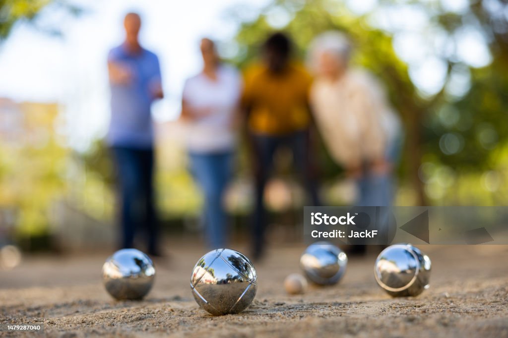 View of petanque shiny silver metal balls laying on the ground outside in the park with group of people standing behind on a sunny day